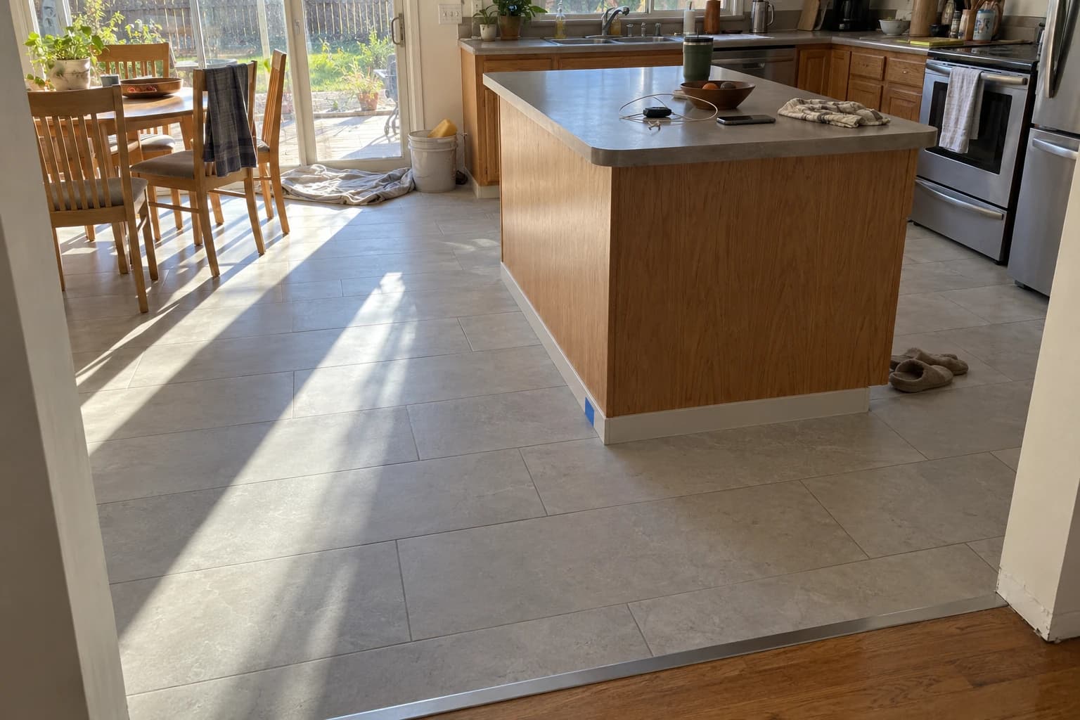 The finished kitchen floor in afternoon light: light gray 12 by 24 inch porcelain laid around the island, running to the dinette table and patio slider, with an aluminum transition strip at the hall opening